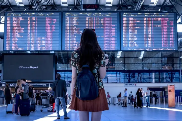한 여성이 가방을 메고 비행기 이착륙시간표를 보고 있는 사진임. 해외여행을 상징함
